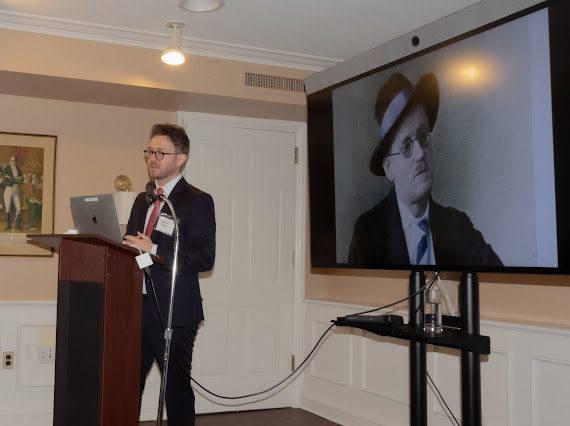 Darren T. Moore en el podio durante la celebración de Bloomsday 2024 de IABANY en la Glucksman Ireland House, con un retrato de James Joyce en pantalla
